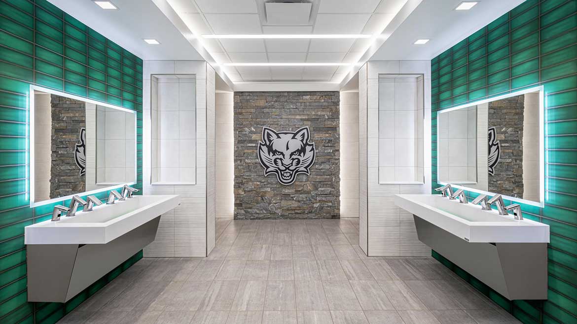 Binghamton University Baseball Stadium Restrooms. Sinks with a row of five faucets, a green wall, lighted mirrors and the baseball logo on the far wall.